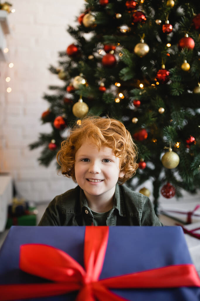 zufriedener Junge mit roten Haaren blickt in die Kamera in der Nähe großer Geschenkschachtel und Weihnachtsbaum auf verschwommenem Hintergrund - Foto, Bild