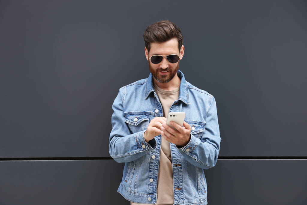 Komea parrakas mies älypuhelimen lähellä harmaa seinä ulkona - Valokuva, kuva