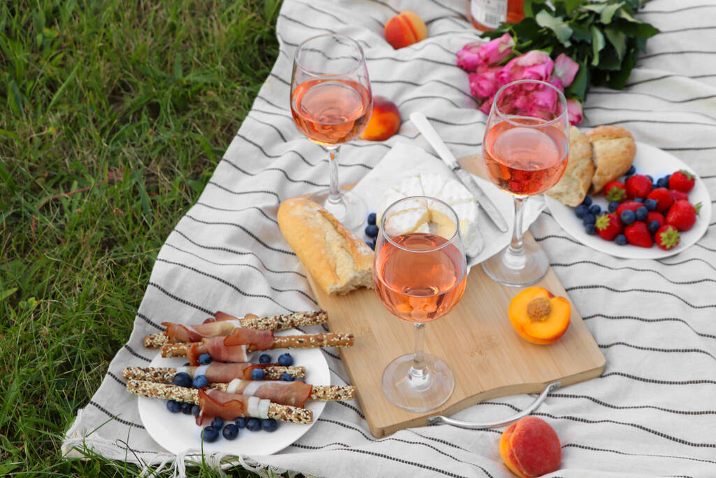 Glasses of delicious rose wine, flowers and food on picnic blanket outdoors - 写真・画像