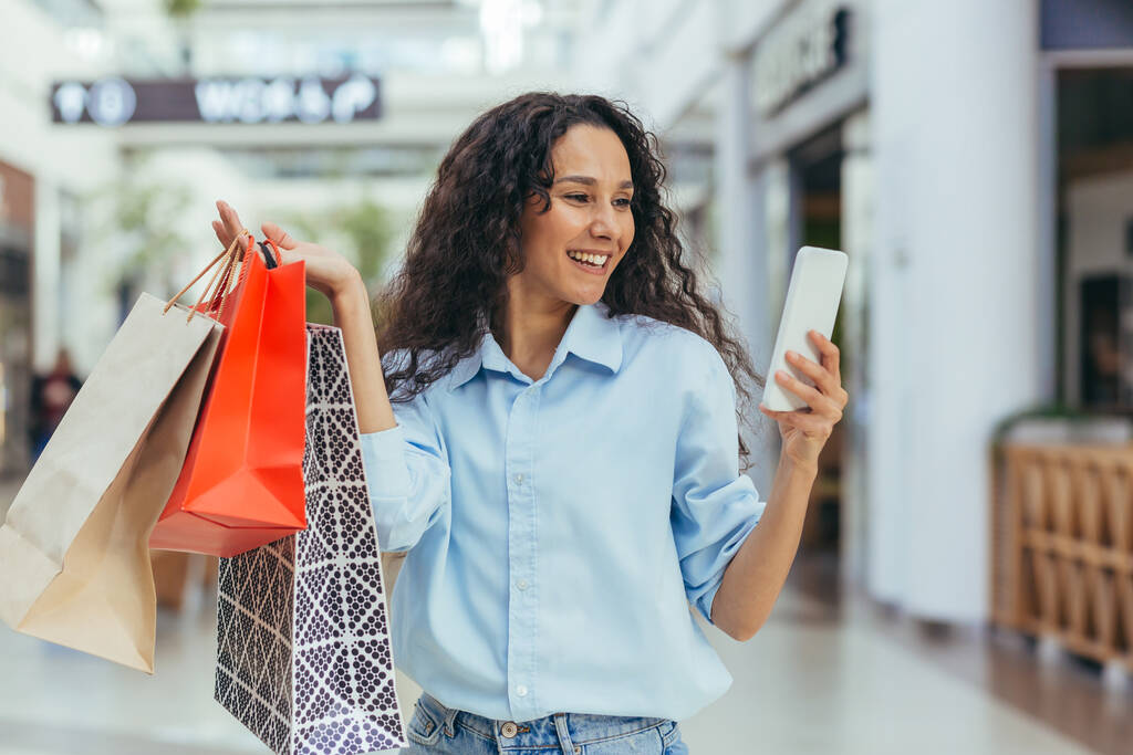 Una atractiva joven latinoamericana está de compras en un centro comercial, sosteniendo bolsas de colores en sus manos. Usa el teléfono, se toma una selfie, sonríe. - Foto, imagen