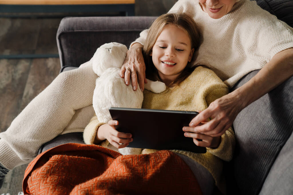 Счастливые бабушка и внучка с помощью планшетного компьютера во время отдыха на диване дома - Фото, изображение