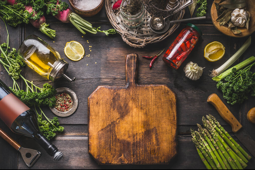 Voedselachtergrond. Diverse ingrediënten en kruiden voor lekker koken rond snijplank op houten rustieke tafel. Olie, citroen, wijnfles, gepekelde chili in pot, asperges, kruiden en specerijen. Kader - Foto, afbeelding