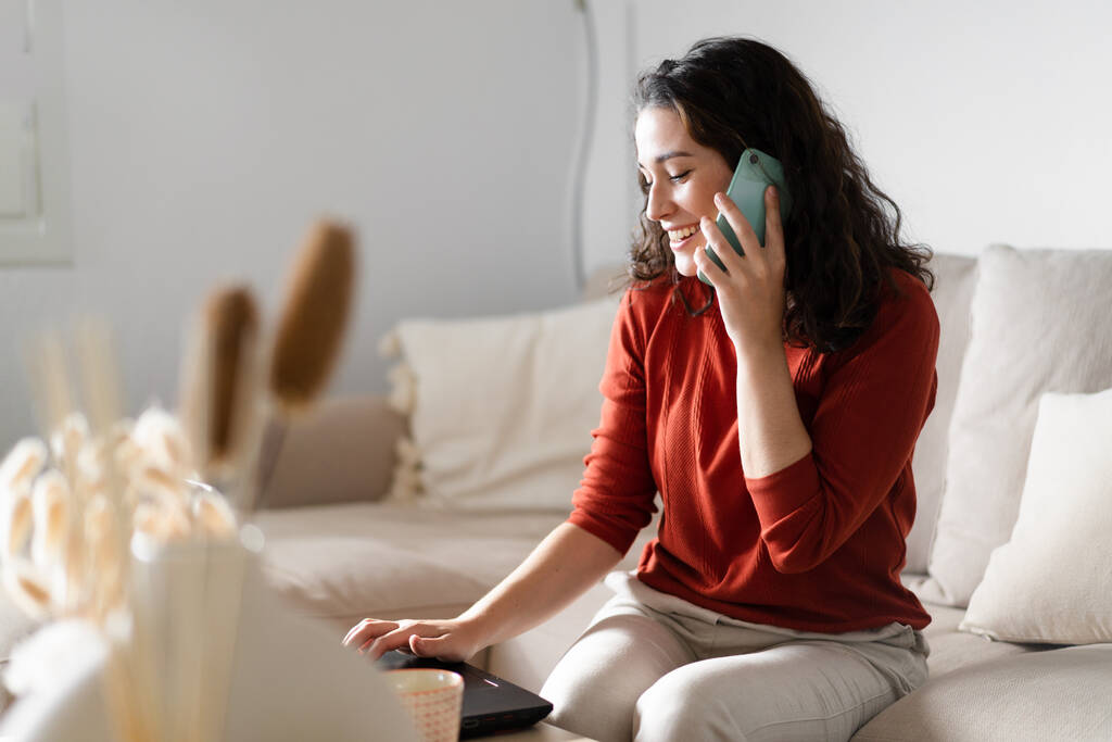 Fiatal boldog nő beszél a telefonon, és gépel a számítógépen ül a kanapén otthon - Fotó, kép