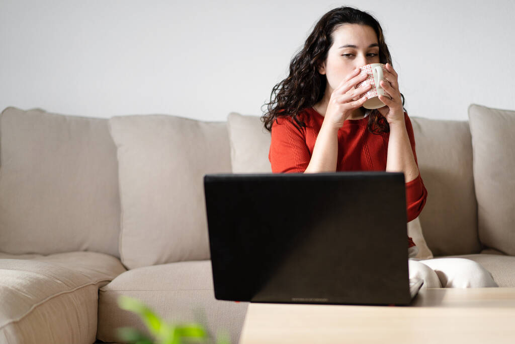 Jonge vrouw die een kopje koffie drinkt en zijdelings naar het computerscherm kijkt terwijl ze thuis op de bank zit. Werken vanuit huis concept - Foto, afbeelding