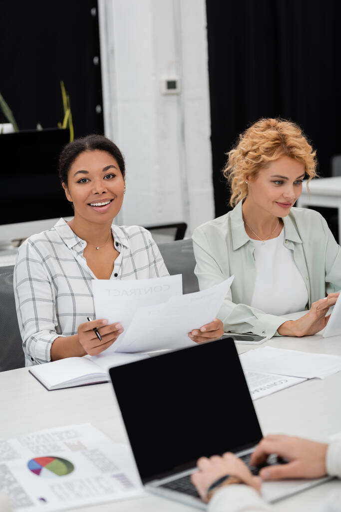 brunette multiracial femme d'affaires tenant des documents près de collègue blonde et ordinateur portable avec écran blanc sur le premier plan flou - Photo, image