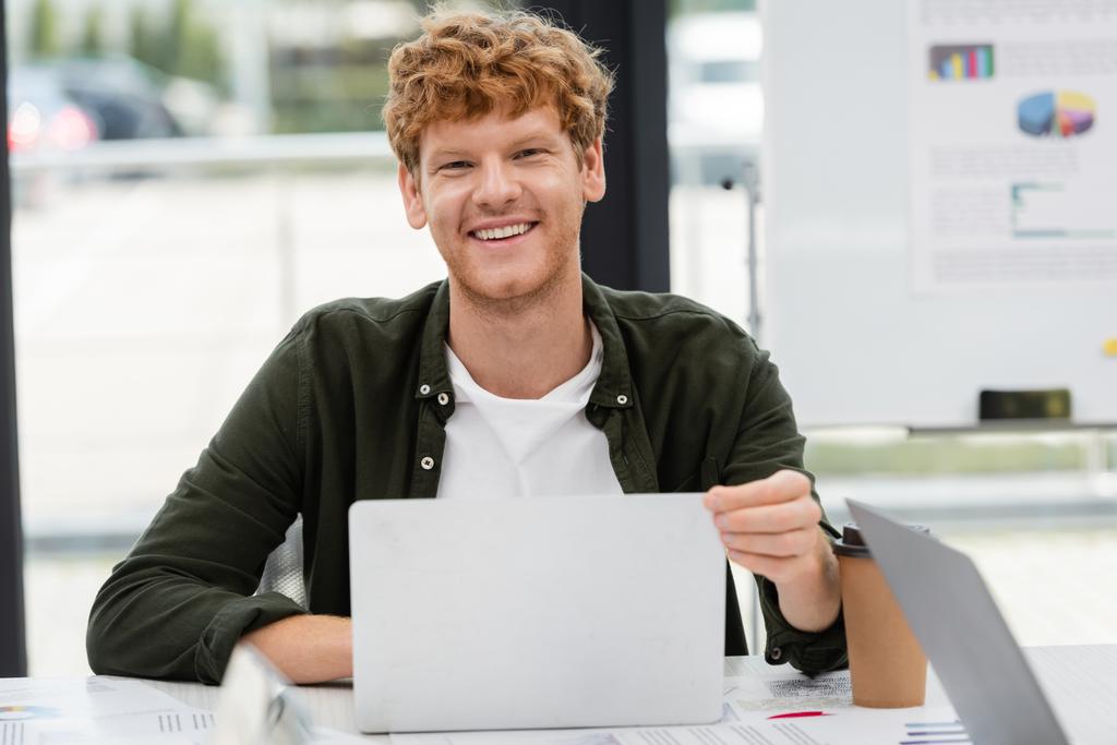 iloinen punapää johtaja hymyilee kameralle lähellä kannettavaa tietokonetta ja paperimukia työpaikalla - Valokuva, kuva