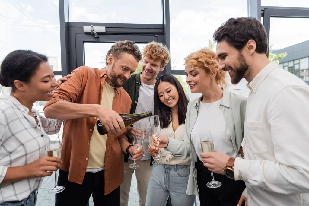 bearded man pouring champagne during corporate party with cheerful multiethnic colleagues  - Photo, Image