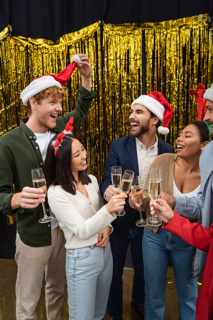 Positiivinen rotujenvälinen liikemiehet katsomalla kollega jouluhattu ja pitämällä samppanjaa virassa  - Valokuva, kuva