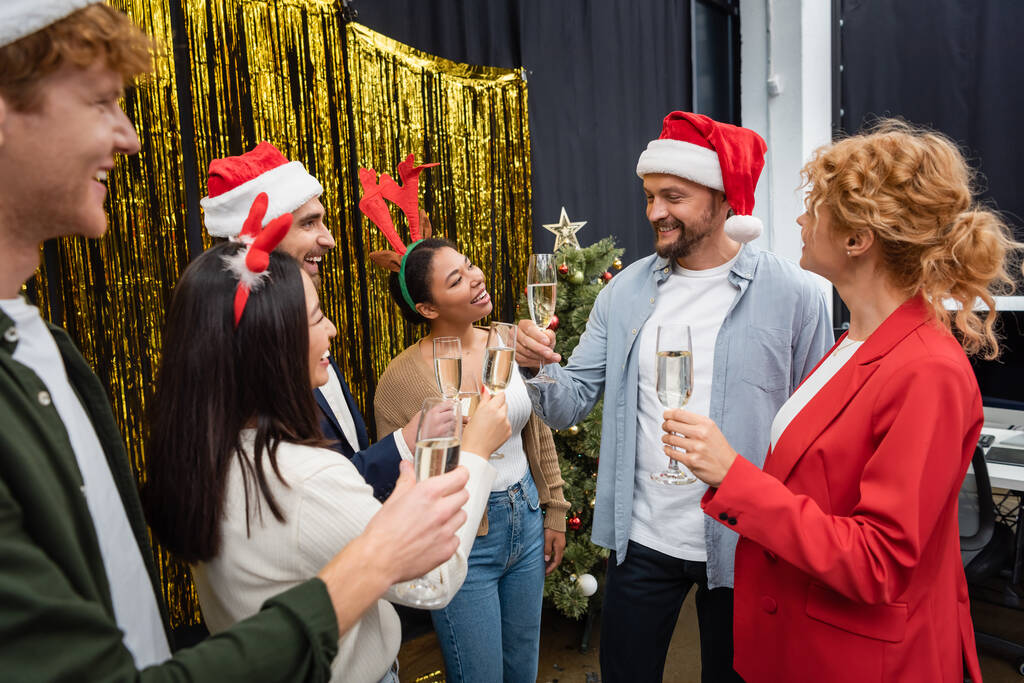 Noel Baba şapkalı gülümseyen işadamı kadehler kaldırıyor ve ofisindeki ırklar arası iş arkadaşlarının yanında şampanya tutuyor.  - Fotoğraf, Görsel