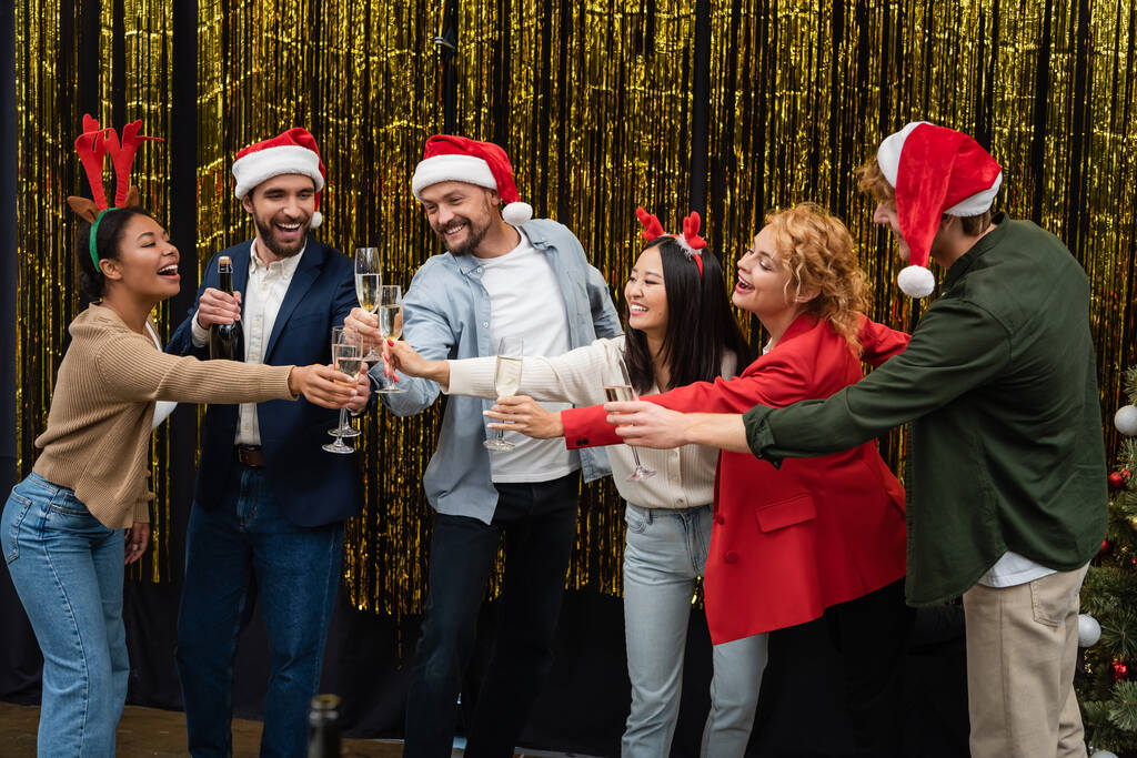 Multikulturelle Geschäftsleute mit weihnachtlichen Stirnbändern klirren neben Lametta im Büro mit Champagner  - Foto, Bild