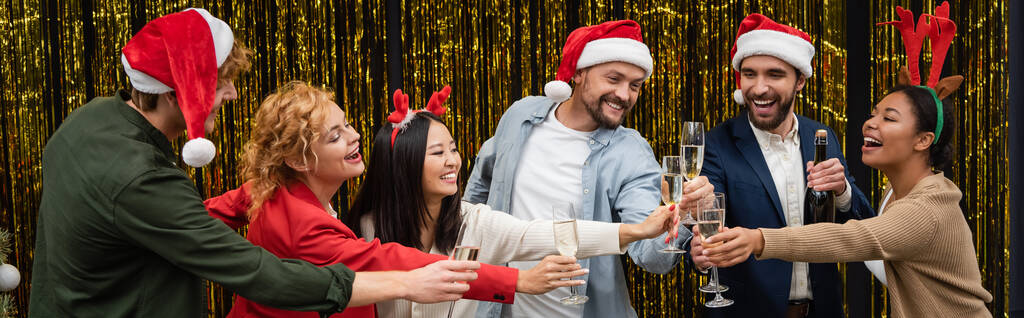 Positive multiethnische Geschäftsleute in Weihnachtsmützen, die mit Kollegen in der Nähe von Lametta Champagner klappern, Banner  - Foto, Bild