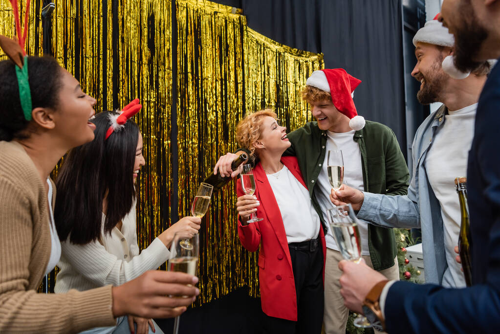 Hymyilevä liikemies kaatamalla samppanjaa lähellä rotujenvälinen kollegansa lasit aikana Christmas Party toimistossa  - Valokuva, kuva