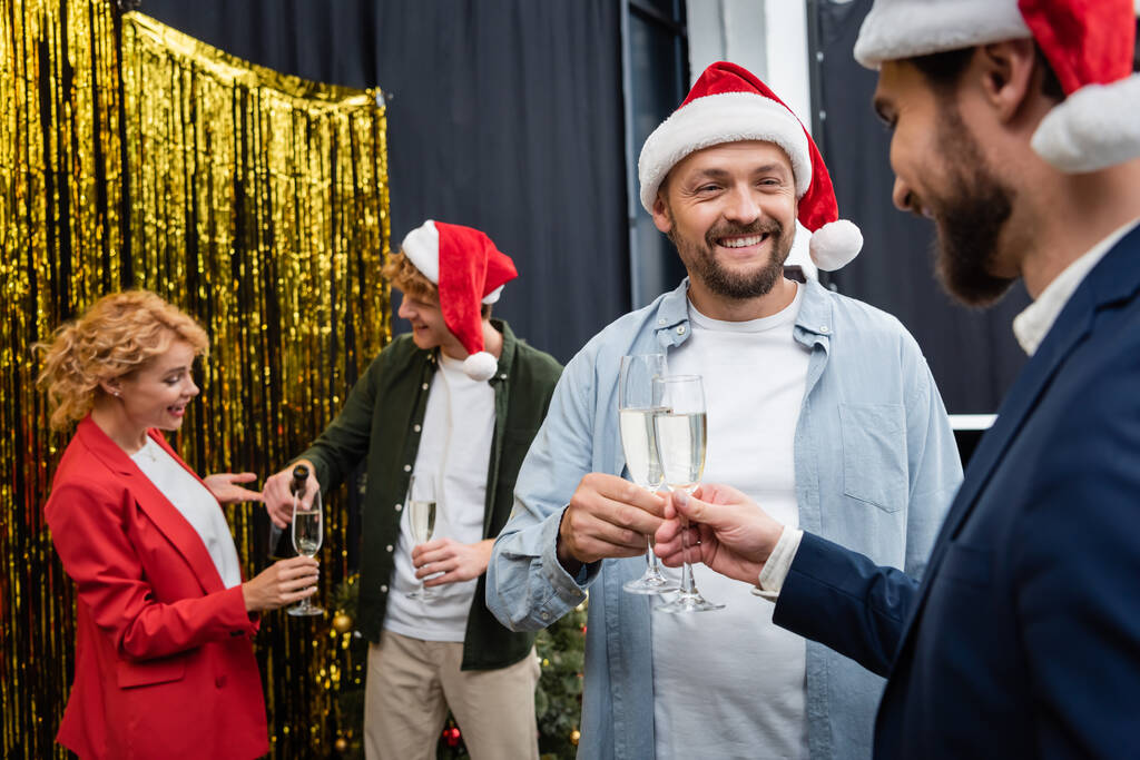 Mosolygó üzletemberek télapó sapkában pezsgőt tartanak homályos kollégák közelében a karácsonyi partin az irodában  - Fotó, kép