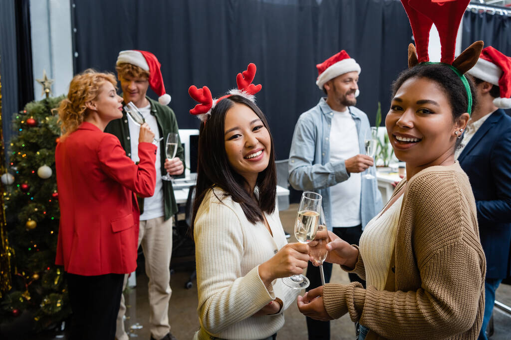 Usmívající se multietnické obchodnice ve vánočních čelenkách držící šampaňské a dívající se na kameru v kanceláři  - Fotografie, Obrázek