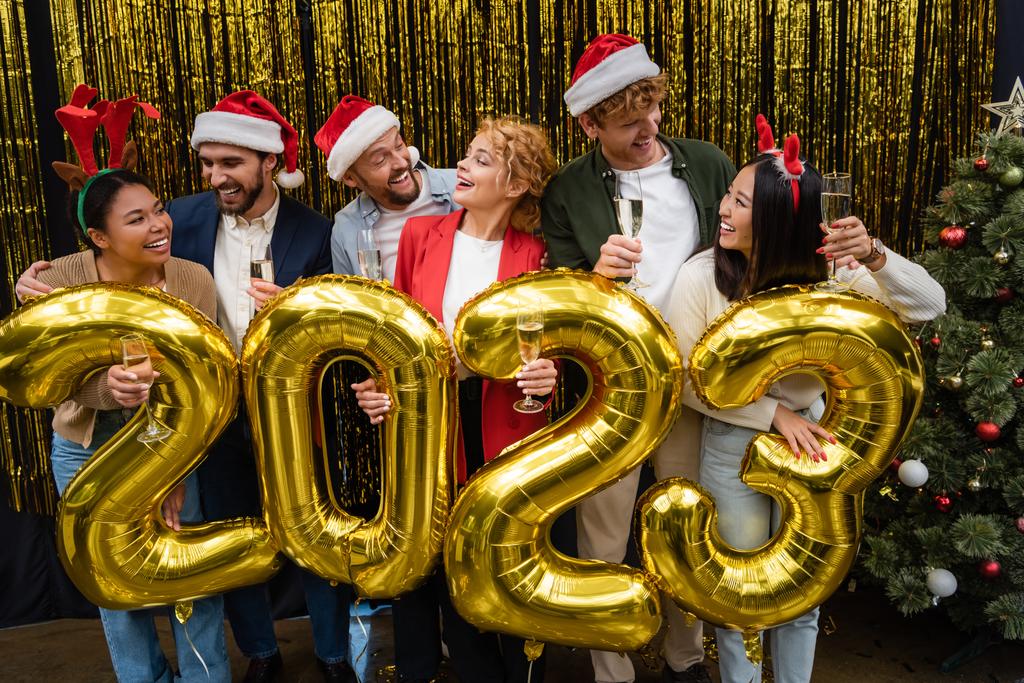 Empresários multiculturais positivos em chapéus de Santa segurando champanhe perto de balões em forma de 2023 no escritório  - Foto, Imagem