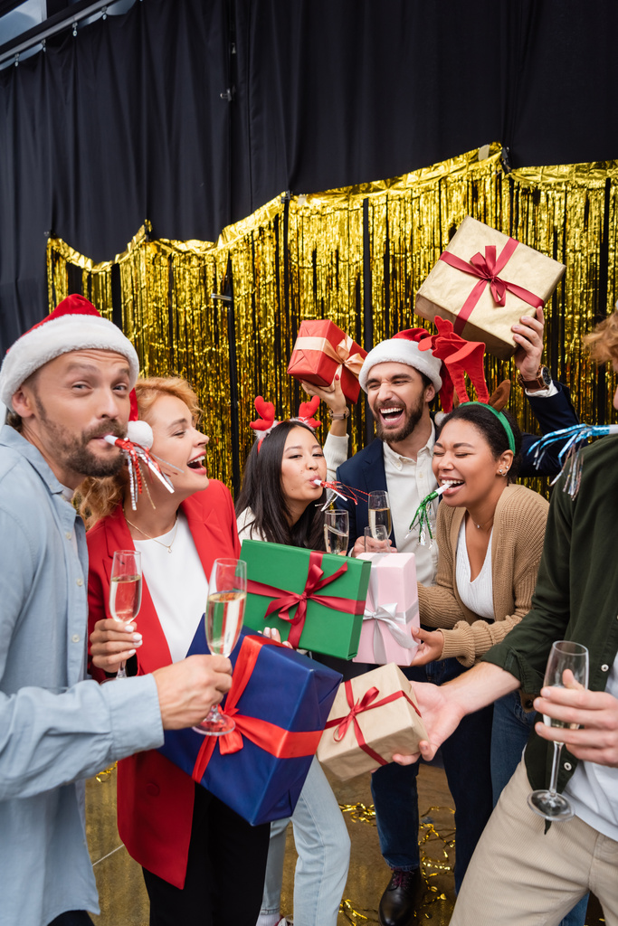 Veselý mezirasoví obchodníci s party rohy drží dárky a šampaňské v kanceláři  - Fotografie, Obrázek