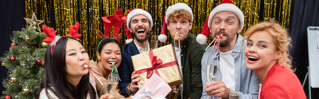 Multiculturele zakenmensen met geschenken en champagne blazen feesthoorns in de buurt van kerstboom in kantoor, banner  - Foto, afbeelding