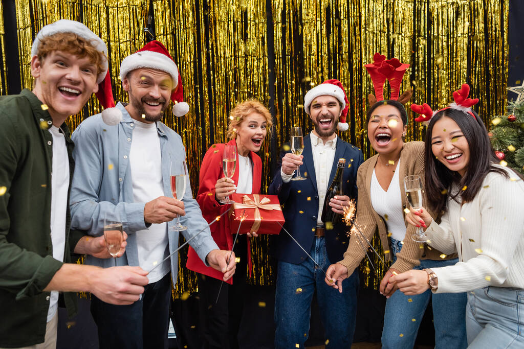 Gente de negocios multiétnicos emocionados celebración de champán y bengalas bajo confeti durante la fiesta de Navidad en la oficina  - Foto, Imagen