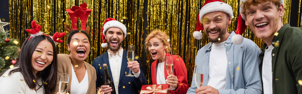 Позитивные межрасовые бизнесмены с бокалами шампанского, стоящие под конфетти во время рождественской вечеринки в офисе, баннер  - Фото, изображение