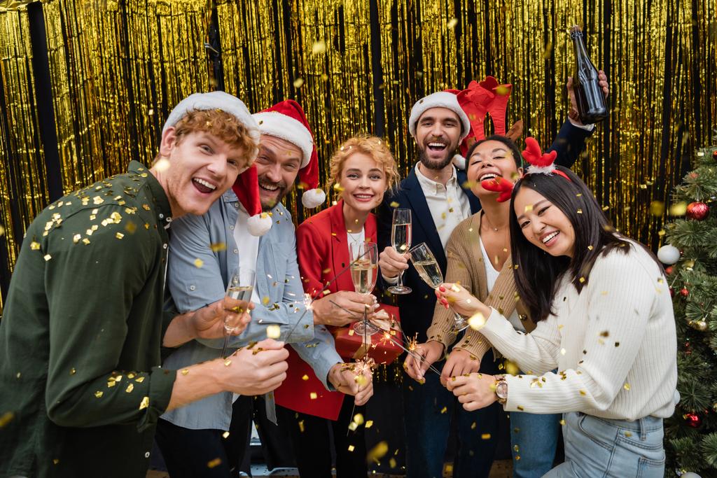 Smiling multiethnic business people in santa hats holding champagne and sparklers under confetti in office  - Photo, Image