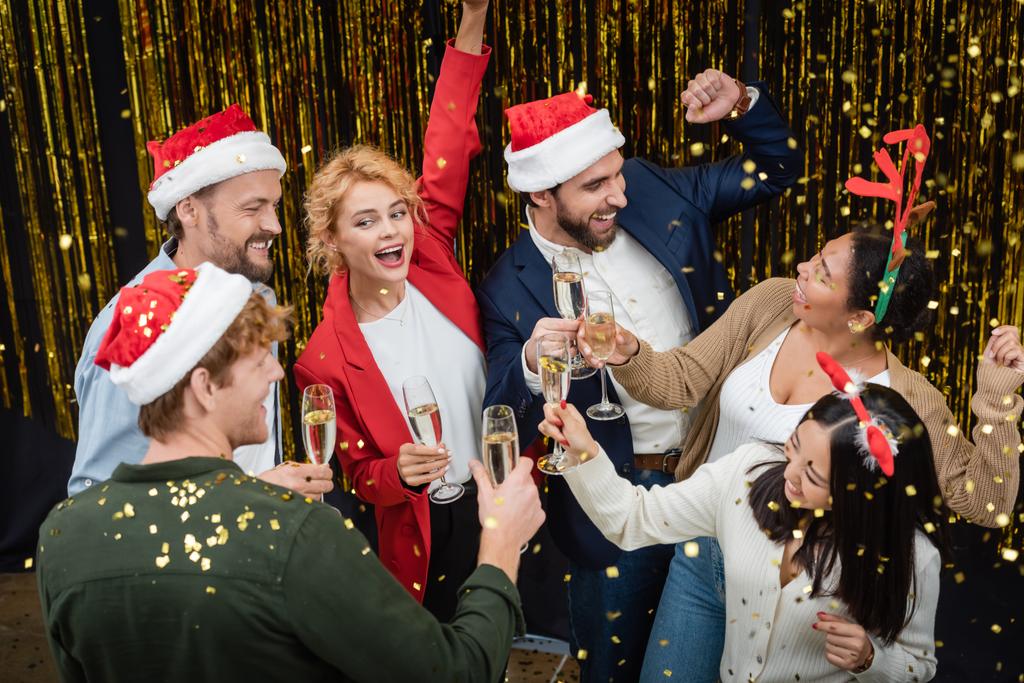 正の異人種間のビジネスの人々がオフィスでクリスマスパーティー中にコンフェッティの下でシャンパンとダンスを保持  - 写真・画像