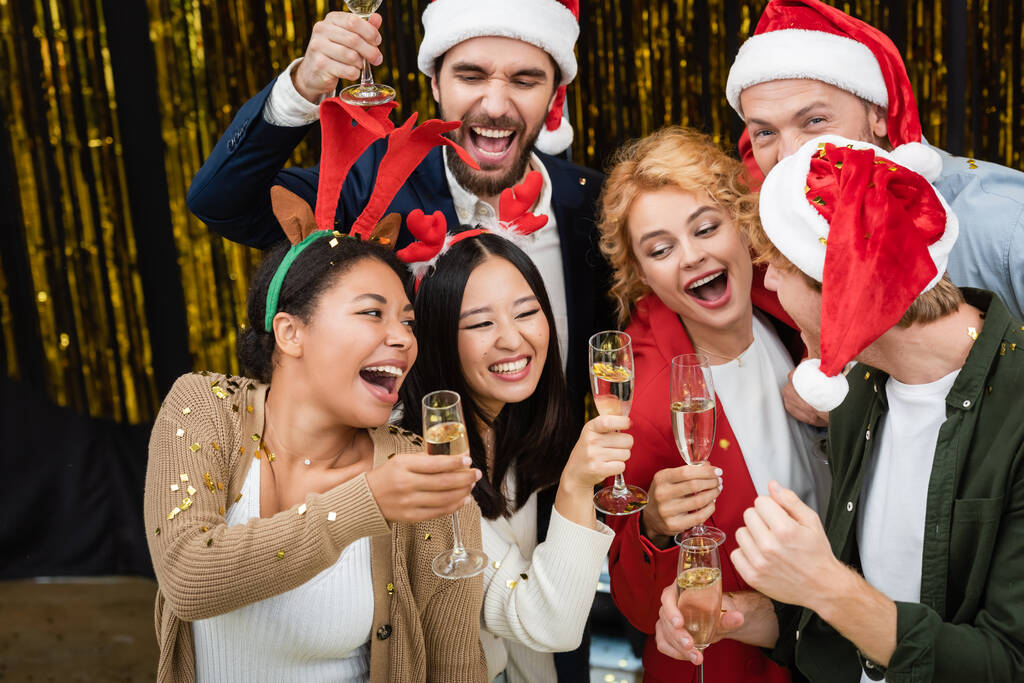 Negócios multiétnicos pessoas segurando champanhe sob confete durante a festa de Natal no escritório  - Foto, Imagem