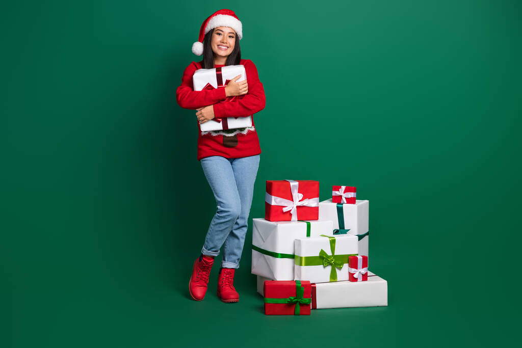 Plná délka fotografie snové okouzlující dívka oblečené ošklivý tisk červená x-mas svetr objímání dárky stack hromádka izolované zelené barvy pozadí. - Fotografie, Obrázek