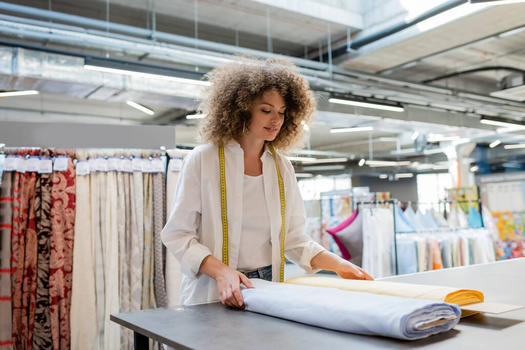 Улыбающаяся продавщица с измерительной лентой, держащая синие и желтые рулоны ткани в текстильном магазине  - Фото, изображение