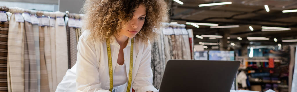 кудрявая продавщица с измерительной лентой с помощью ноутбука в текстильном магазине, баннер  - Фото, изображение