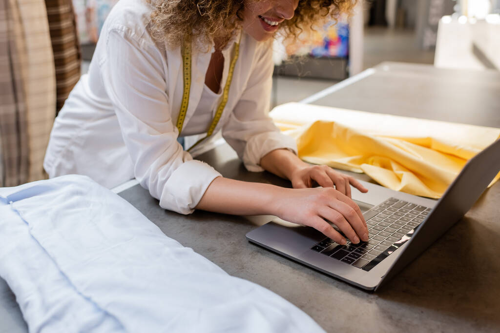 abgeschnittene Ansicht der lockigen Verkäuferin lächelnd und mit Laptop in der Nähe von Stoffrollen im Textilgeschäft  - Foto, Bild