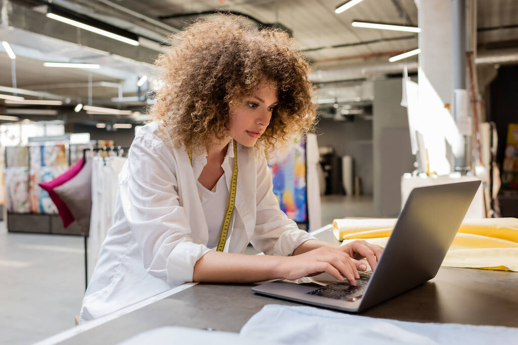 curly and young saleswoman using laptop while working in textile shop  - Photo, Image