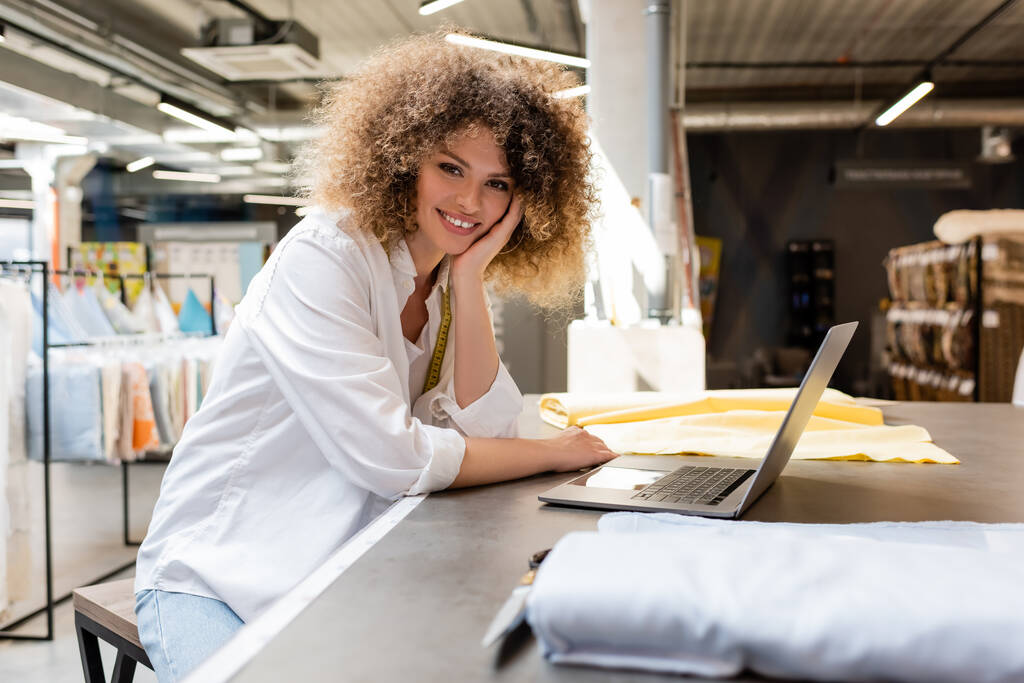 Lockige Verkäuferin mit Maßband lächelt neben Laptop und Stoffrollen im Textilgeschäft  - Foto, Bild