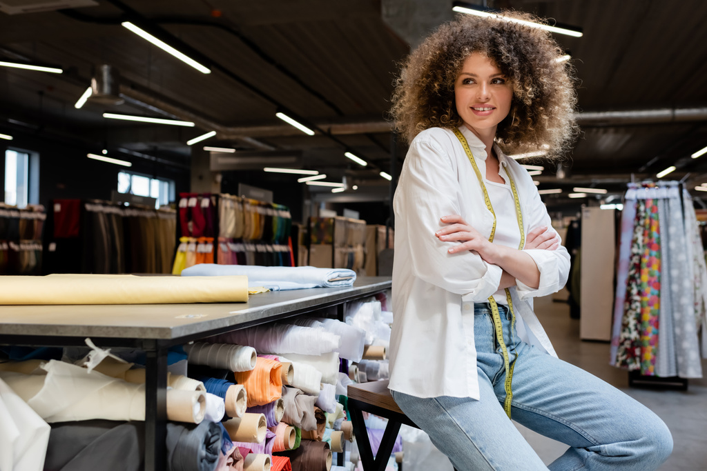 joyful saleswoman with curly hair posing with crossed arms near rack with fabric rolls in textile shop  - Photo, Image