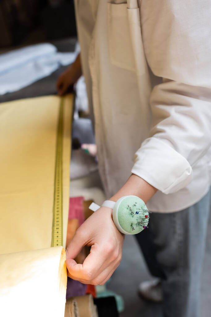 Teilansicht einer Verkäuferin mit Nadelkissen auf der Hand, die gelben Stoff im Textilgeschäft misst  - Foto, Bild