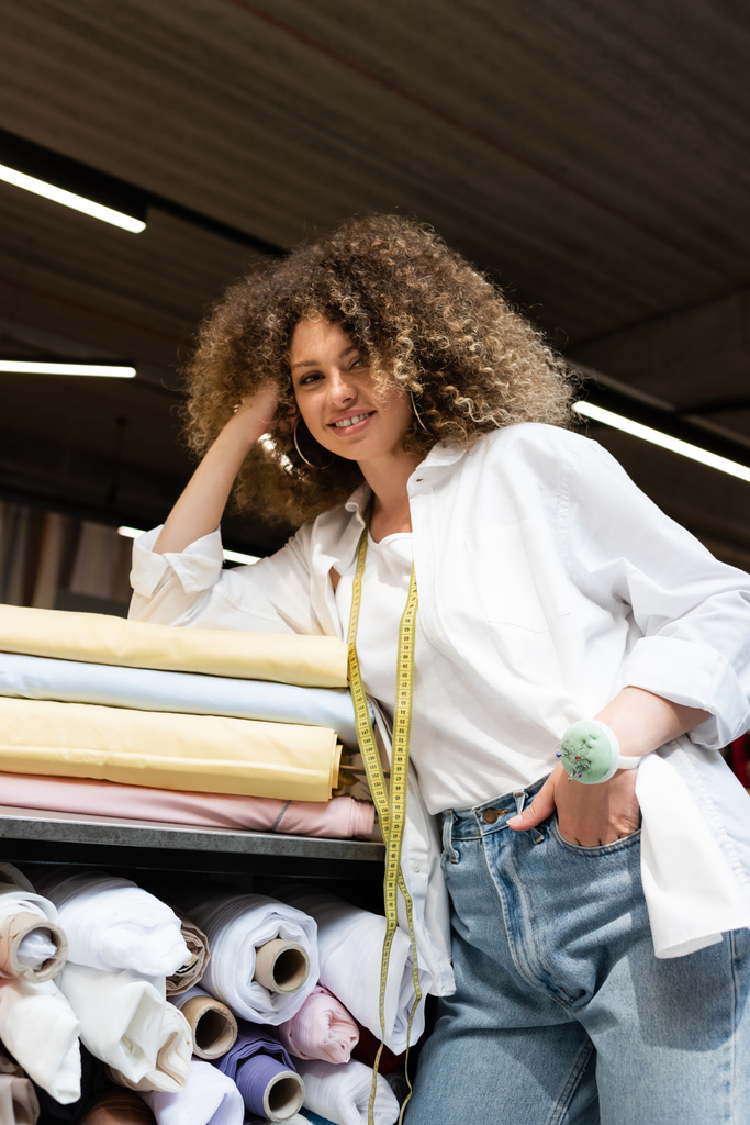 улыбающаяся продавщица с игольчатой подушкой, позирующая с рукой в кармане, опираясь на стойку с разноцветными рулонами ткани  - Фото, изображение