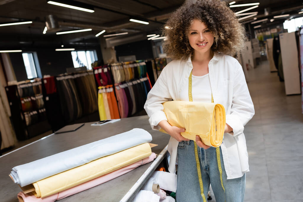 положительная продавщица держит желтый пастельный рулон ткани в текстильном магазине  - Фото, изображение
