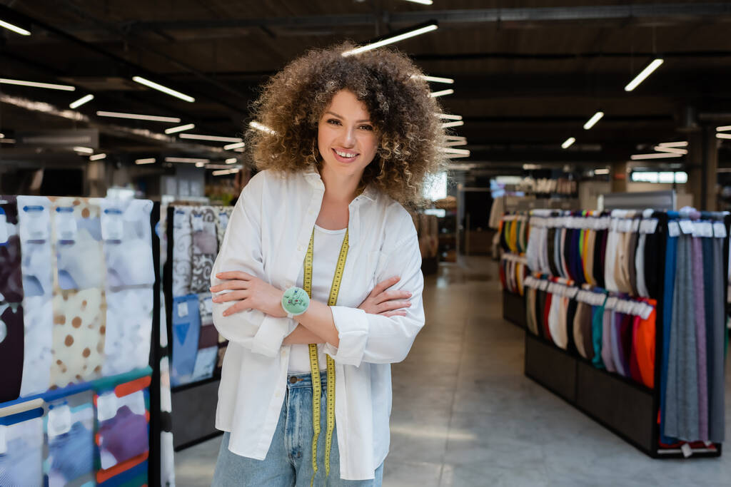 vendedora feliz y rizado de pie con los brazos cruzados cerca de filas borrosas de diversos tejidos en la tienda textil - Foto, imagen