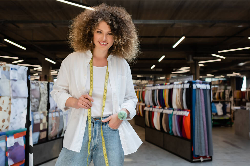 kręcone sprzedawczyni z taśmą pomiarową i poduszką igły stojącej z ręką w kieszeni dżinsów w sklepie włókienniczym - Zdjęcie, obraz