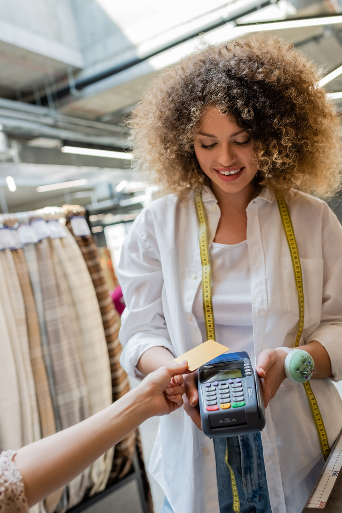 vendedor feliz e encaracolado com fita métrica segurando terminal de pagamento perto do cliente com cartão de crédito na loja têxtil - Foto, Imagem