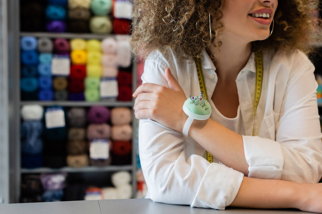 partial view of smiling saleswoman with measuring tape and needle cushion near counter and blurred rack with yarn skeins - Photo, Image