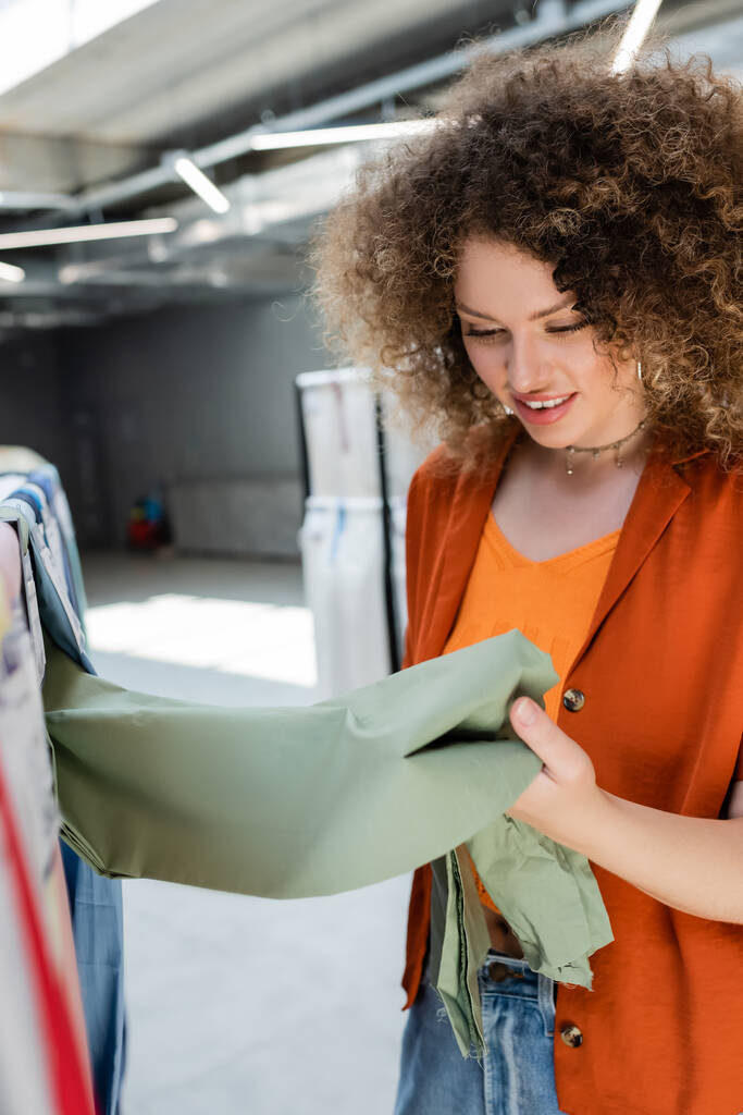 curly woman smiling while choosing fabric in textile shop - Photo, Image