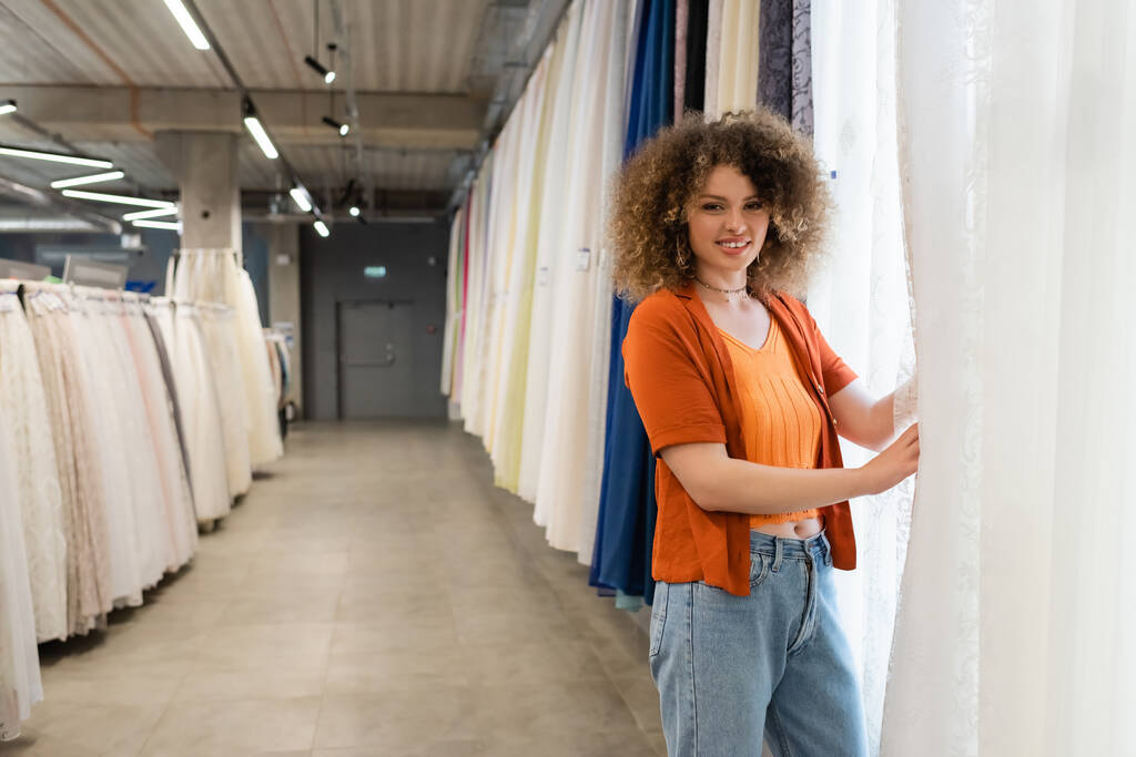 mulher feliz e encaracolado olhando para a câmera perto variedade de cortinas na loja têxtil - Foto, Imagem