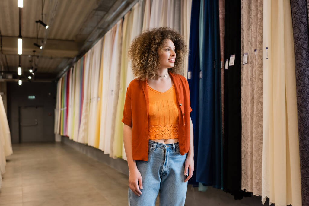 улыбающаяся женщина с вьющимися волосами, смотрящая на разноцветные занавески в текстильном магазине - Фото, изображение