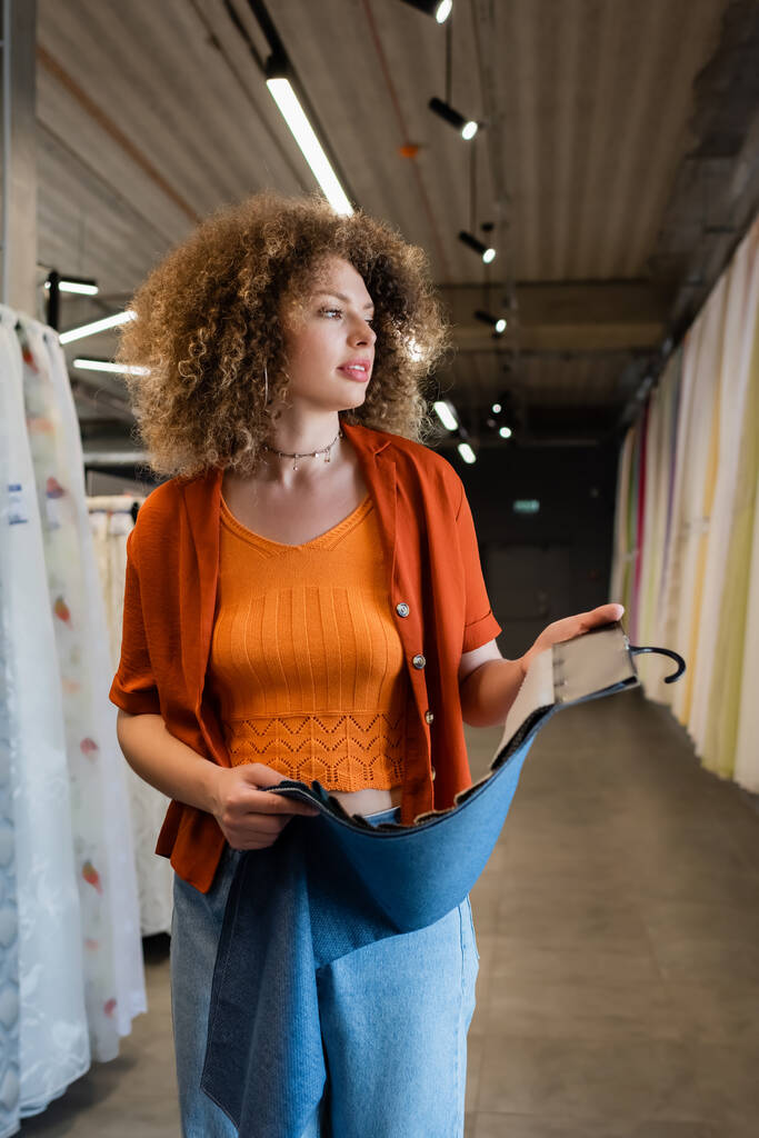 Giovane donna riccia che tiene campioni di stoffa e distoglie lo sguardo nel negozio di tessuti  - Foto, immagini
