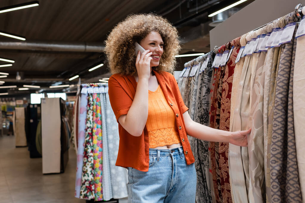 Usmívající se kudrnatá žena mluví na smartphone a dotýká tkaniny v textilním obchodě  - Fotografie, Obrázek
