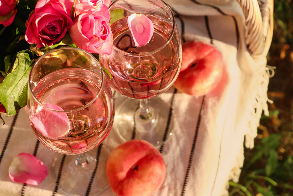 Skleničky lahodného růžového vína s okvětními lístky, květinami a broskvemi na bílé piknikové přikrývce venku, nad okny - Fotografie, Obrázek