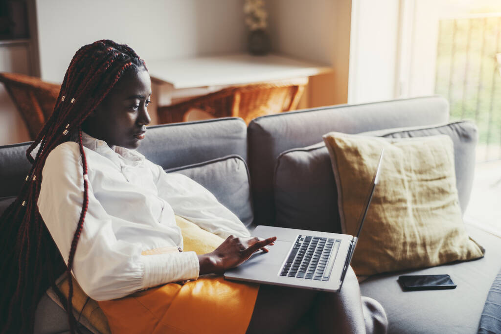 Nézet egy figyelmes fiatal fekete nő vállalkozó fehér ingben ül a kanapén a háztartási nappali és egy netbook; fekete nő használja a laptop a kanapén, körülvéve párnák - Fotó, kép