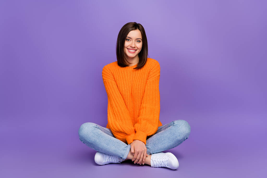 Täysikokoinen kuva upea nuori tyttö istuu lattialla ristissä jalat pukeutunut tyylikäs neulottu oranssi näyttää eristetty violetti väri tausta. - Valokuva, kuva