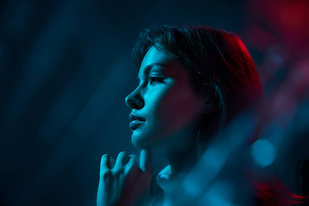 Portrét mladé krásné ženy pózující izolované přes modré pozadí studia v neonovém světle. Fotografie z boku. Koncept kultury mládeže, emocí, výrazu obličeje, módy. Kopírovat prostor pro inzerci - Fotografie, Obrázek
