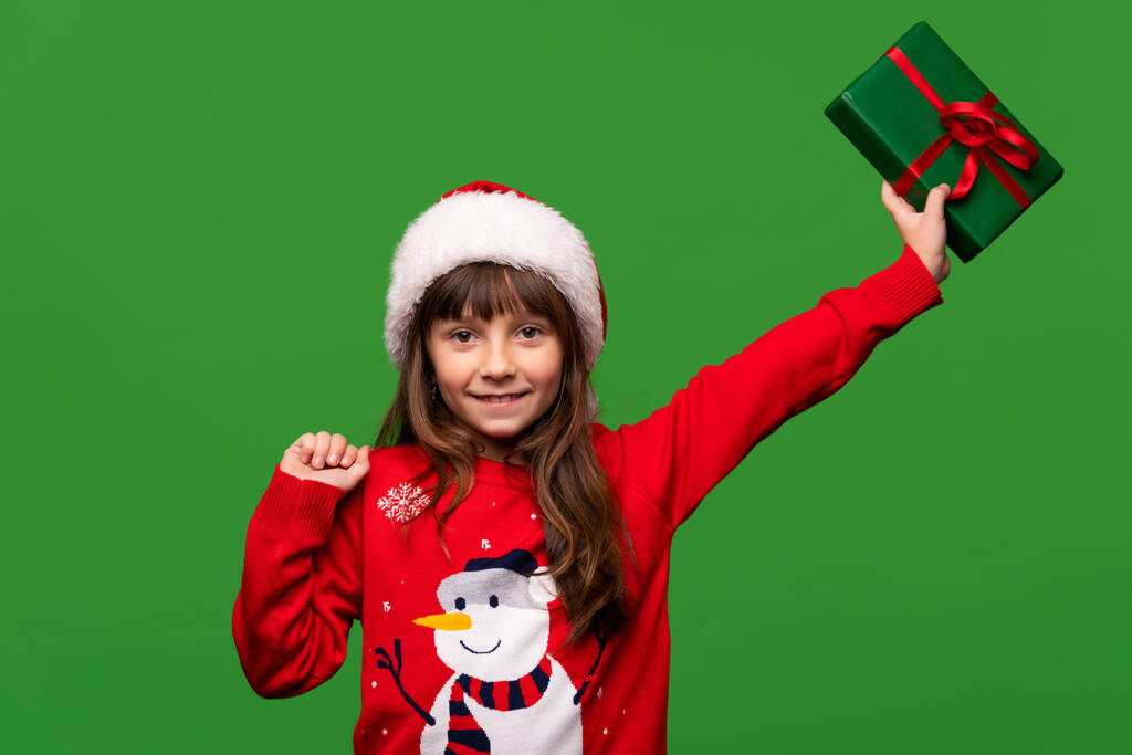 Улыбающаяся маленькая девочка в шляпе Санты показывает прекрасный завернутый подарок. С Новым 2023 годом и Рождеством Христовым. Портрет изолирован на зеленом фоне. Высокое качество фото - Фото, изображение